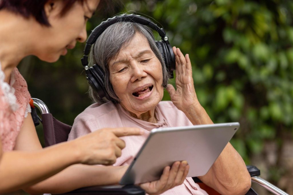 Senior mental health patient wears headphones while looking at tablet held by aid